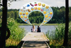 Zwei Frauen am See, über ihnen eine Sprechblase mit Emojis wie Totenkopf und Aubergine.